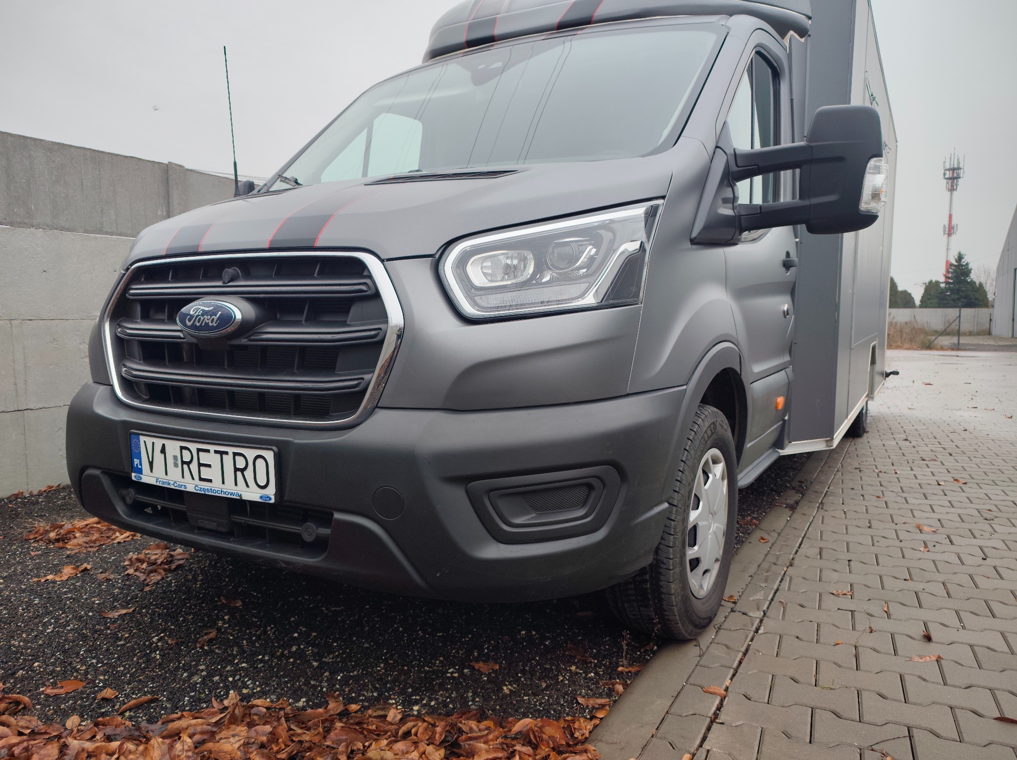 Szary Ford Transit z czarnymi i czerwonymi pasami, tablica rejestracyjna V1-RETRO, zaparkowany na kostce brukowej z liśćmi. Widok z przodu, pochmurny dzień.