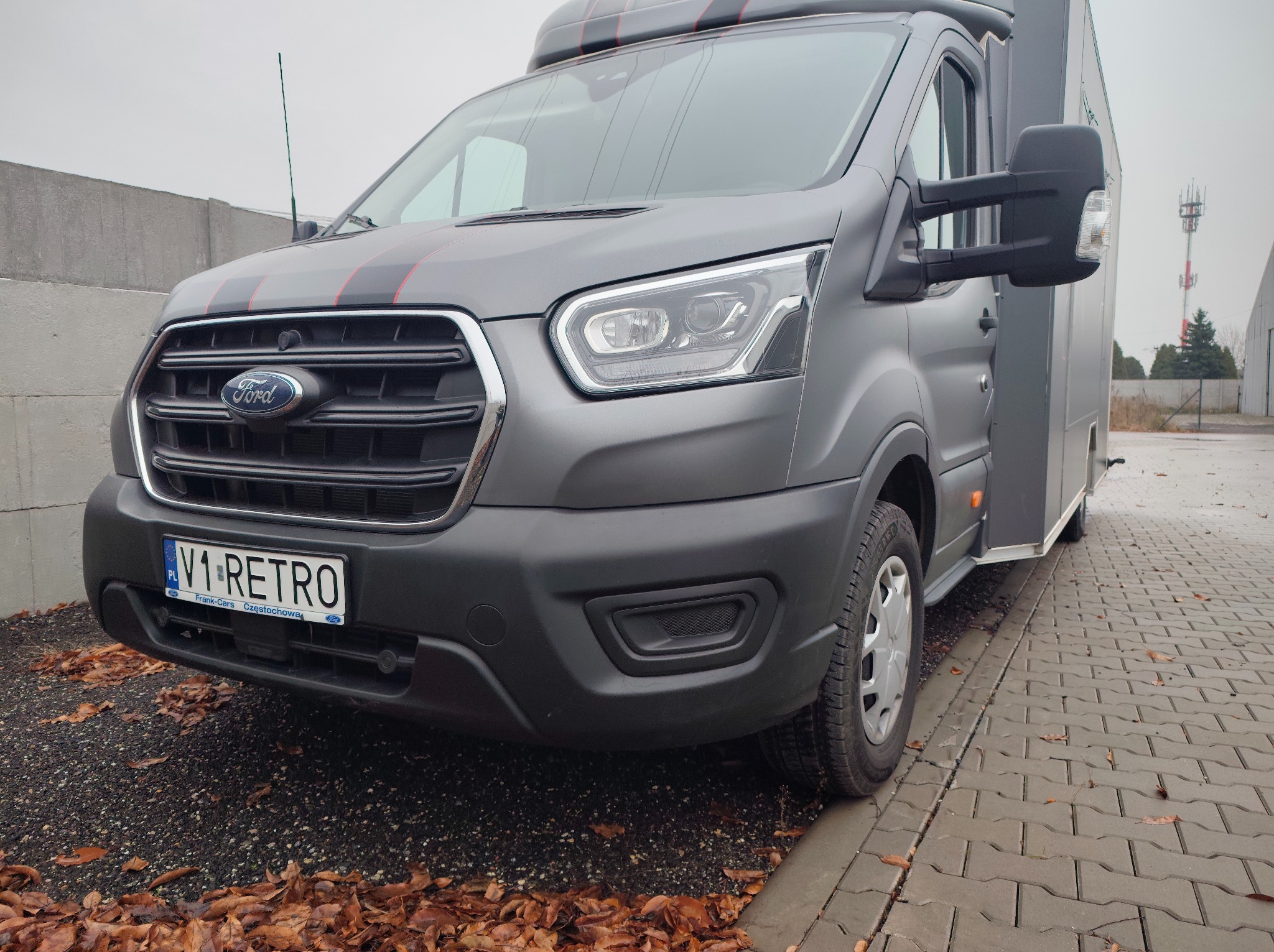 Szary Ford Transit z czerwonymi pasami, tablica V1-RETRO. Matowa folia na karoserii, widok z przodu na tle szarej kostki brukowej i jesiennych liści.