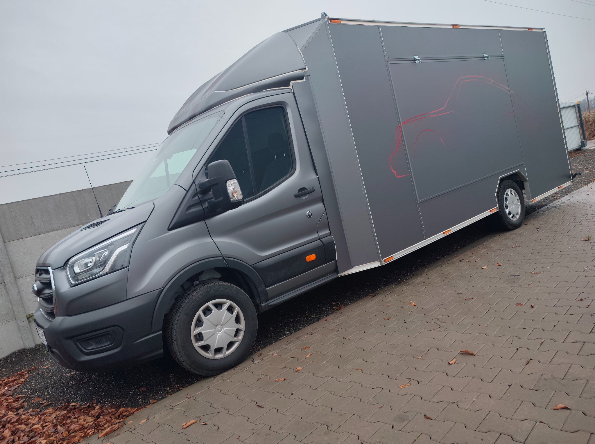 Szara furgonetka z przyczepą, na której widnieje czerwony zarys samochodu. Pojazd stoi na kostce brukowej, w tle szary mur i pochmurne niebo.