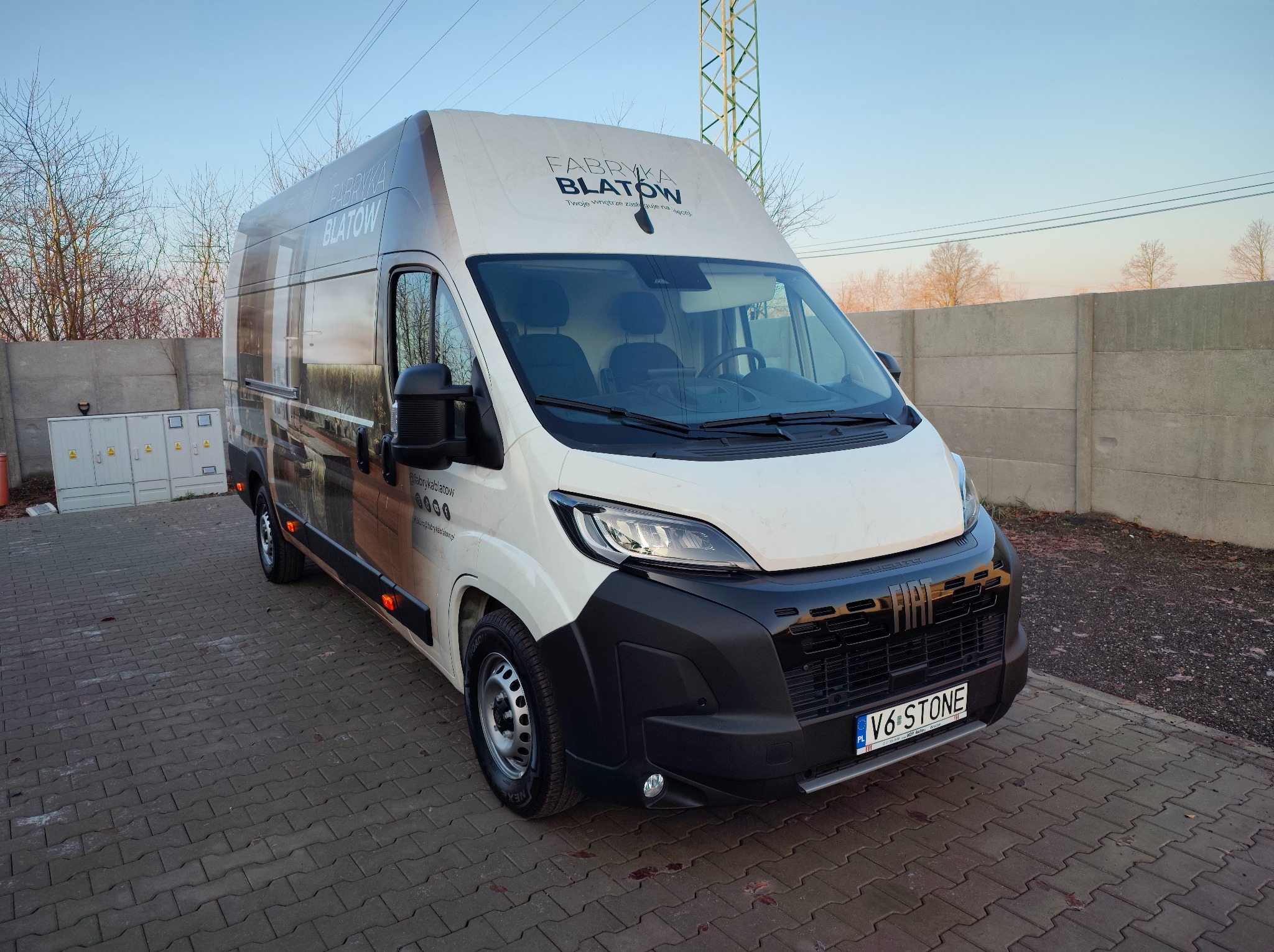 Biały bus z grafiką reklamową firmy Fabryka Blatów, zaparkowany na kostce brukowej. Widoczny bok pojazdu z logo i danymi kontaktowymi. W tle betonowy mur i słup energetyczny.