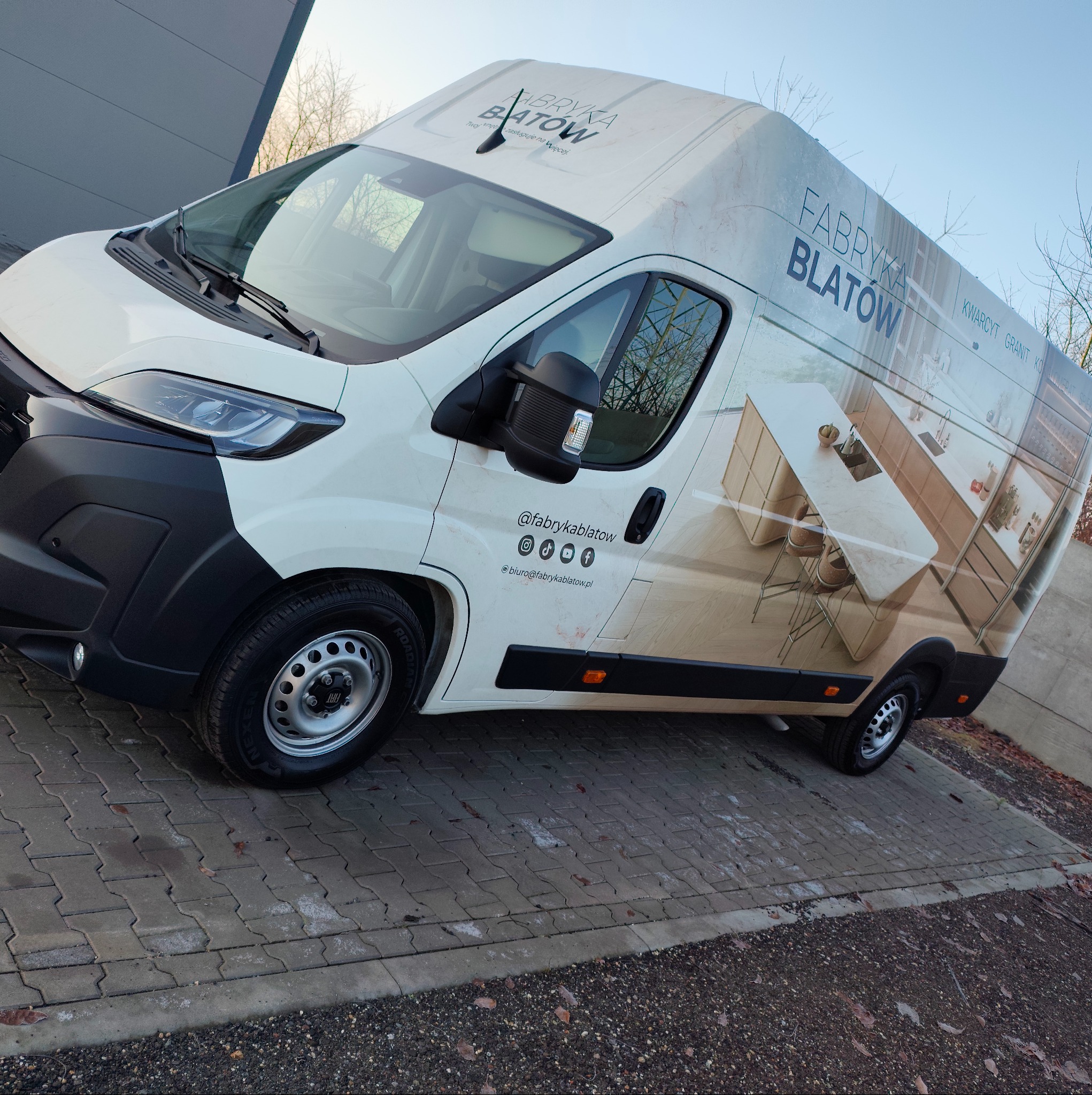 Oklejony van 'Fabryka Blatów' z grafiką blatu kuchennego i danymi kontaktowymi, zaparkowany na kostce brukowej. Reklama mobilna firmy.