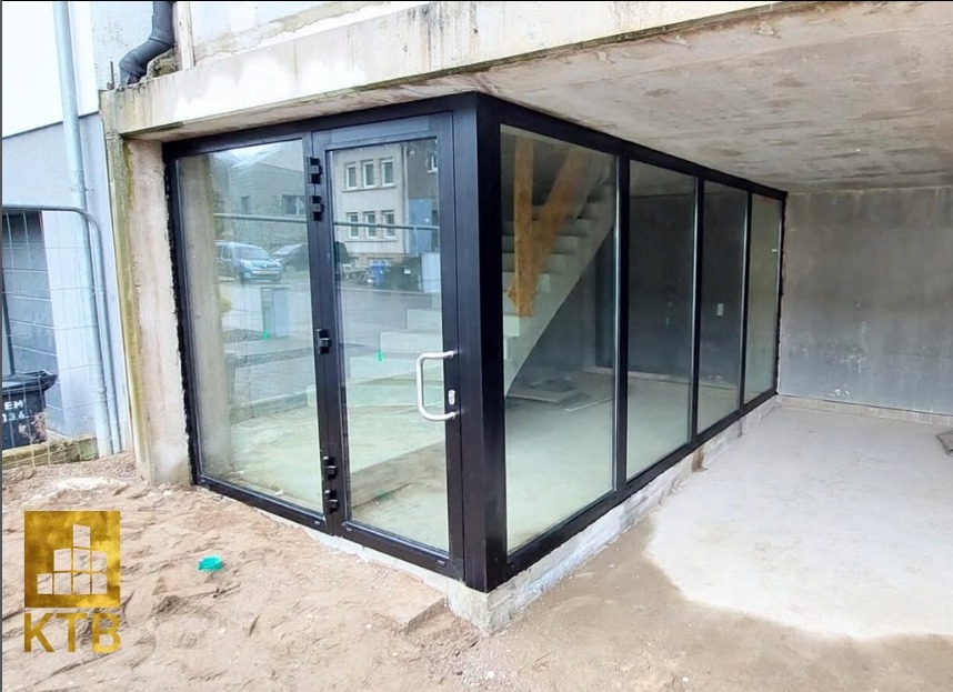 Nowoczesne, czarne drzwi przesuwne z dużymi szybami, w surowym wnętrzu z widocznymi schodami. Na zewnątrz piasek i budynki w tle. Minimalistyczny design.