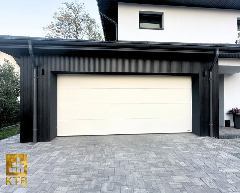 Nowoczesna brama garażowa segmentowa w kolorze białym, zamontowana w czarnej, drewnianej obudowie garażu przylegającego do białego domu. Kostka brukowa przed garażem.
