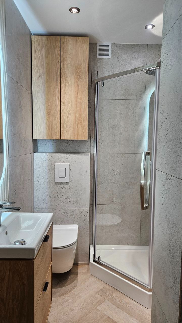 Nowoczesna łazienka po remoncie: szare płytki, kabina prysznicowa ze szklanymi drzwiami, drewniana szafka pod umywalką i podwieszana toaleta. Jasne, punktowe oświetlenie.