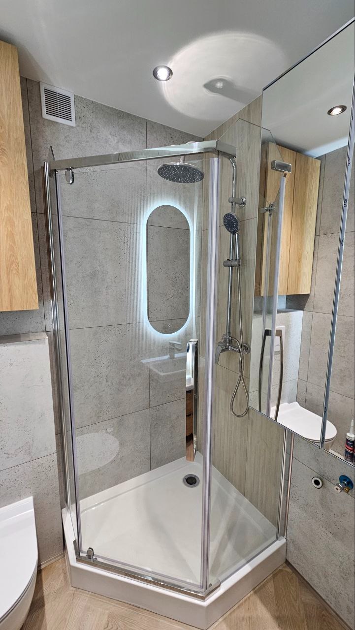 Nowoczesna kabina prysznicowa z deszczownicą i podświetlanym lustrem w odnowionej łazience, wykończona szarą płytką imitującą beton i drewnianą szafką.