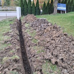 ELEKTROSOLUTIONS KAMIL JUDKA - Wykopany rów w ziemi na trawniku, prawdopodobnie pod kable elektryczne. W tle widoczny biały słup i tablica informacyjna firmy. Jesienny krajobraz w tle.