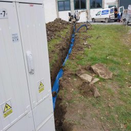 ELEKTROSOLUTIONS KAMIL JUDKA - Wykopany rów z niebieską rurą osłonową, obok biała szafa elektryczna z ostrzeżeniami. W tle koparka i samochód z napisem Automatyka Bram.