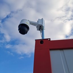 ELEKTROSOLUTIONS KAMIL JUDKA - Kamera przemysłowa PTZ na maszcie, widok z dołu na tle błękitnego nieba z chmurami. Obudowa kamery biała, maszt srebrny, fragment czerwonej konstrukcji w dolnej części kadru.