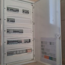 ELEKTROSOLUTIONS KAMIL JUDKA - Otwarta, biała skrzynka rozdzielcza z bezpiecznikami i schematem, zamontowana na surowej ścianie. Widoczne oznaczenia obwodów i wyłączniki.