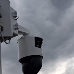 ELEKTROSOLUTIONS KAMIL JUDKA - Kamera przemysłowa Hikvision na słupie, skierowana w górę na tle pochmurnego nieba. Widoczny uchwyt montażowy i obudowa kamery w kolorze biało-czarnym.