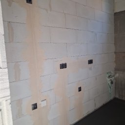 ELEKTROSOLUTIONS KAMIL JUDKA - Ściana z betonu komórkowego z zamontowanymi puszkami elektrycznymi i częściowo położoną gładzią. Widoczne przewody w rogu pomieszczenia.