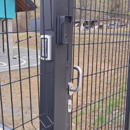 ELEKTROSOLUTIONS KAMIL JUDKA - Nowoczesna, czarna brama posesyjna z panelem dotykowym, wideodomofonem i metalowym uchwytem. W tle widoczny dom z niebieskim akcentem i zalesione wzgórze.