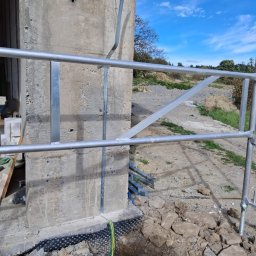 ELEKTROSOLUTIONS KAMIL JUDKA - Betonowy słup z zamontowaną metalową konstrukcją, widoczny kabel w żółto-zielonej izolacji, ziemia i niebo w tle. Prace budowlane w toku.