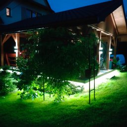 ELEKTROSOLUTIONS KAMIL JUDKA - Oświetlona altana ogrodowa nocą, z podświetlonymi roślinami i elementami konstrukcyjnymi, tworząca nastrojowy klimat. Widoczne detale drewnianej konstrukcji i zielony trawnik.