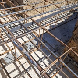 ELEKTROSOLUTIONS KAMIL JUDKA - Zbrojenie fundamentów z widocznymi spawami i elementami stalowymi, przygotowane do zalania betonem. Widok z góry na siatkę zbrojeniową w wykopie.