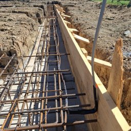 ELEKTROSOLUTIONS KAMIL JUDKA - Zbrojenie fundamentów z wyprowadzonym przewodem elektrycznym w szalunku drewnianym. Widok na wykop, w tle dom w budowie i zalesione wzgórza.