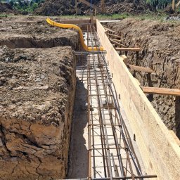 ELEKTROSOLUTIONS KAMIL JUDKA - Zbrojenie fundamentów w wykopie z drewnianym szalunkiem, widoczna żółta rura instalacyjna. W tle góra ziemi i zalesione wzgórza w słoneczny dzień.