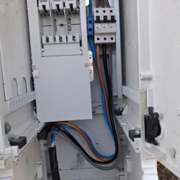 ELEKTROSOLUTIONS KAMIL JUDKA - Otwarta skrzynka elektryczna z widocznymi bezpiecznikami, przewodami w kolorach: brązowym, niebieskim i czarnym. Widoczne elementy montażowe.