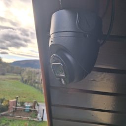 ELEKTROSOLUTIONS KAMIL JUDKA - Kamera kopułkowa CCTV zamontowana na drewnianej konstrukcji, z widokiem na zielony krajobraz. Czarna obudowa kamery kontrastuje z jasnym tłem.