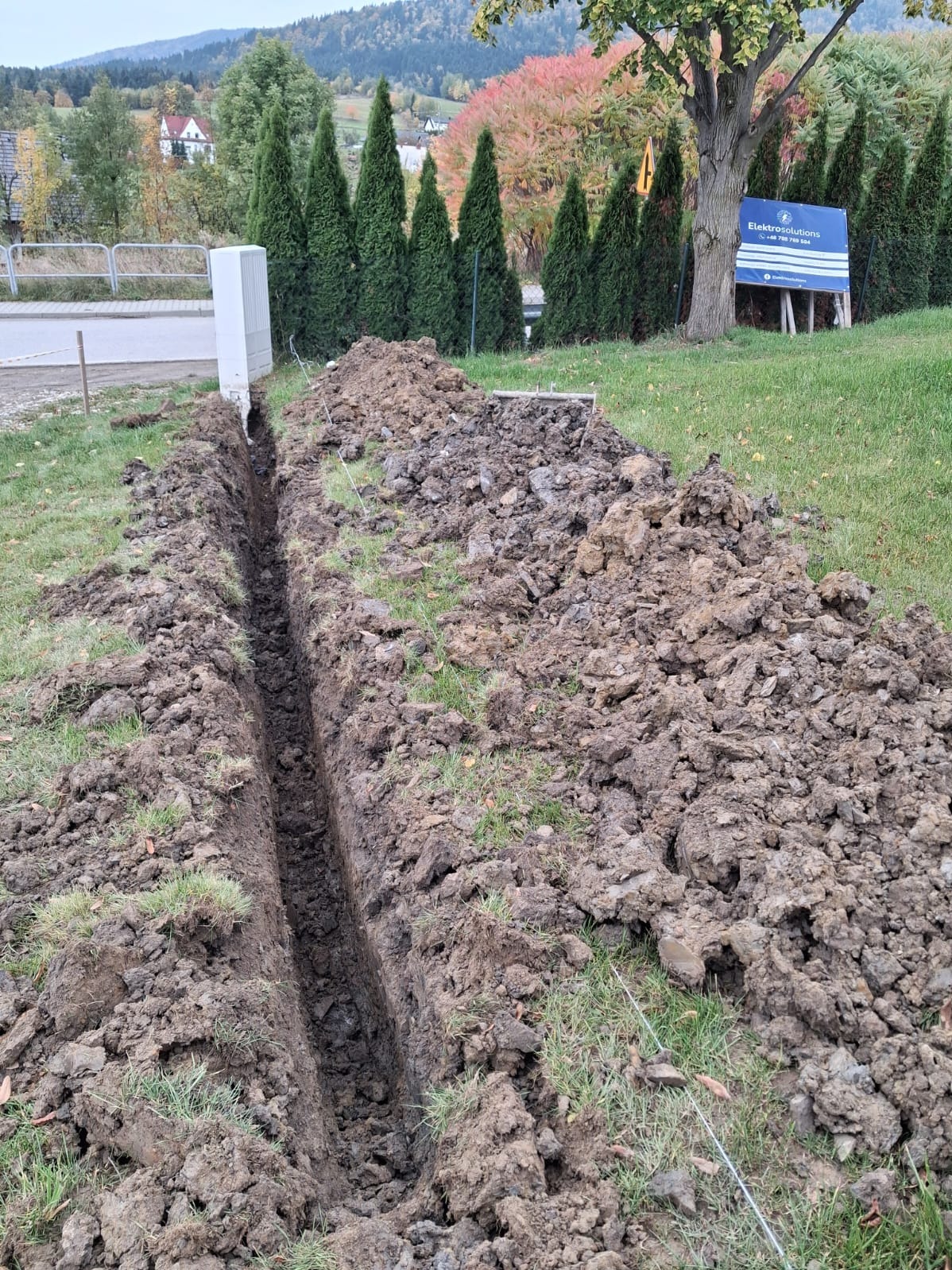 Wykopany rów w ziemi na trawniku, prawdopodobnie pod kable elektryczne. W tle widoczny biały słup i tablica informacyjna firmy. Jesienny krajobraz w tle.