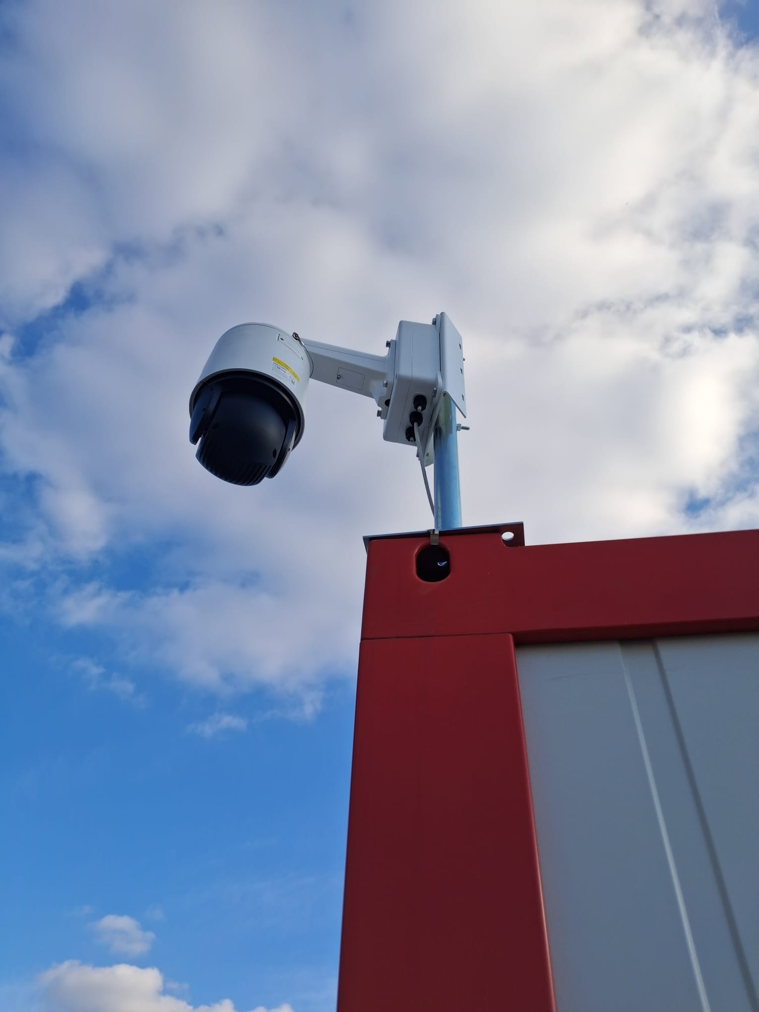 Kamera przemysłowa PTZ na maszcie, widok z dołu na tle błękitnego nieba z chmurami. Obudowa kamery biała, maszt srebrny, fragment czerwonej konstrukcji w dolnej części kadru.