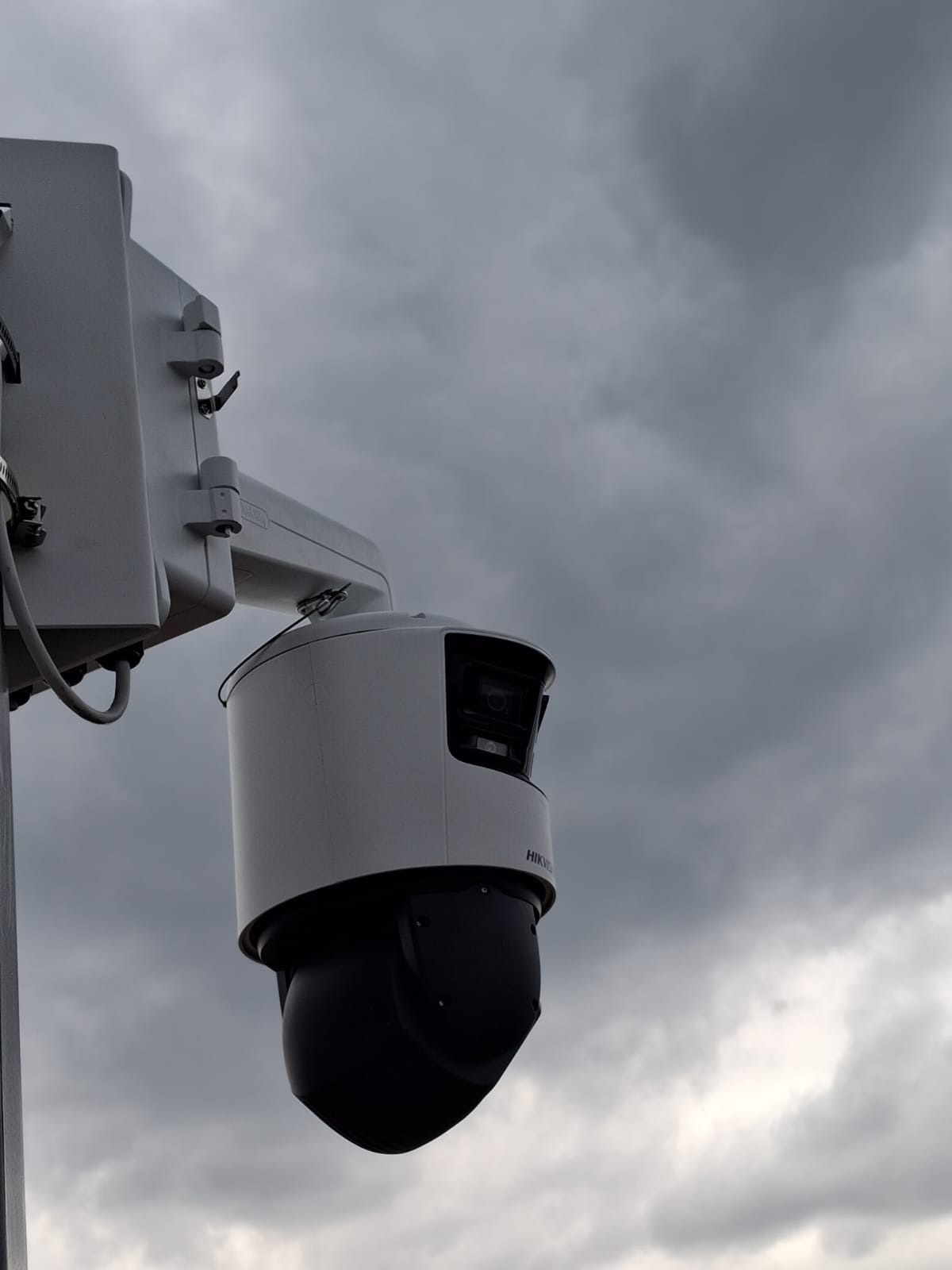 Kamera przemysłowa Hikvision na słupie, skierowana w górę na tle pochmurnego nieba. Widoczny uchwyt montażowy i obudowa kamery w kolorze biało-czarnym.