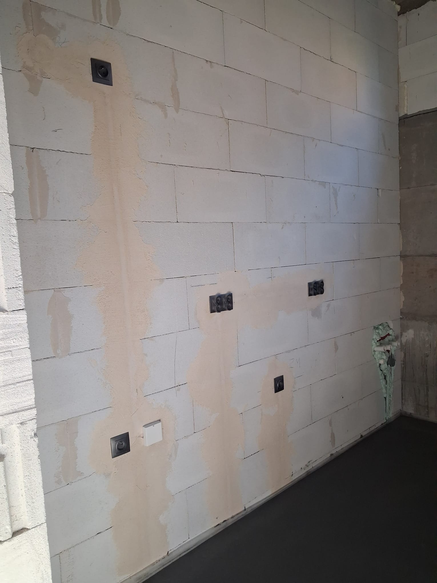 Ściana z betonu komórkowego z zamontowanymi puszkami elektrycznymi i częściowo położoną gładzią. Widoczne przewody w rogu pomieszczenia.