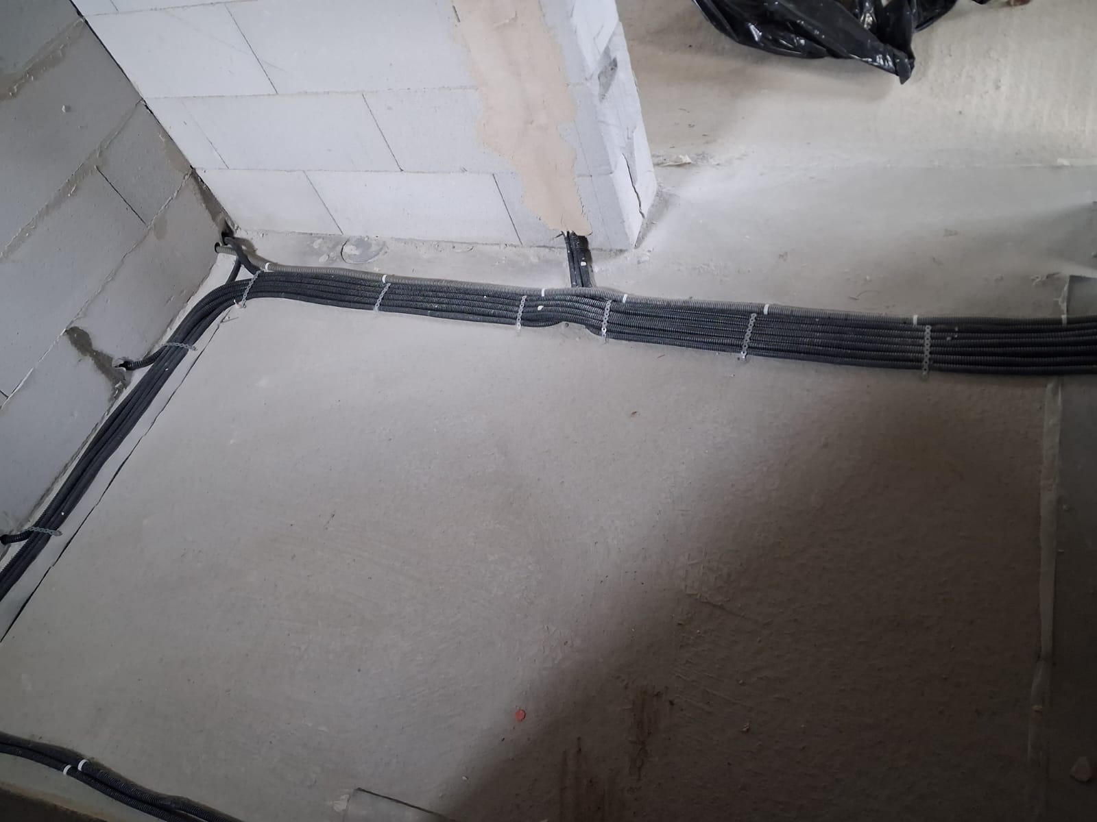 Szereg czarnych, karbowanych rur instalacyjnych ułożonych równolegle na szarej, betonowej podłodze w surowym wnętrzu, przymocowanych białymi opaskami zaciskowymi.