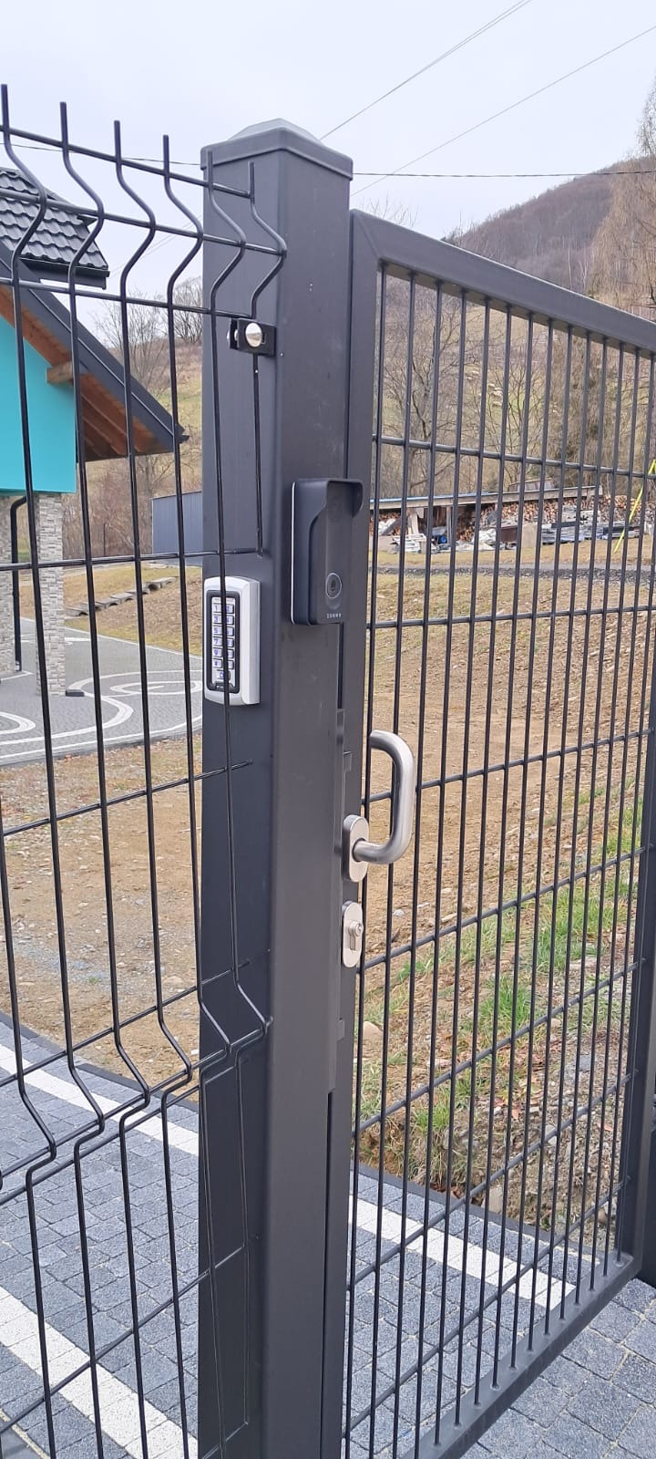 Nowoczesna, czarna brama posesyjna z panelem dotykowym, wideodomofonem i metalowym uchwytem. W tle widoczny dom z niebieskim akcentem i zalesione wzgórze.