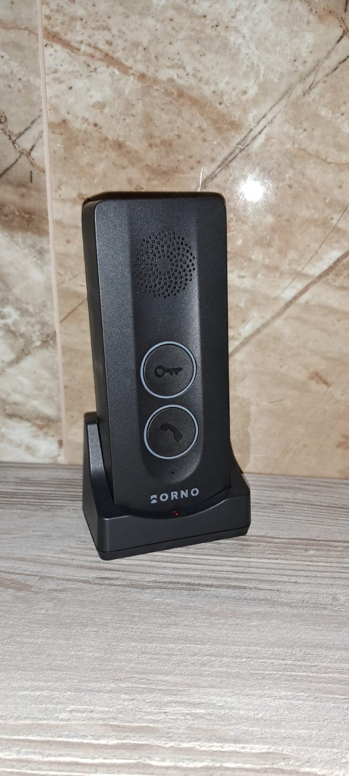 Czarny, nowoczesny wideodomofon ZORNO na podstawce, z przyciskami klucza i słuchawki, na tle marmurowej ściany i drewnianego blatu. Minimalistyczny design.