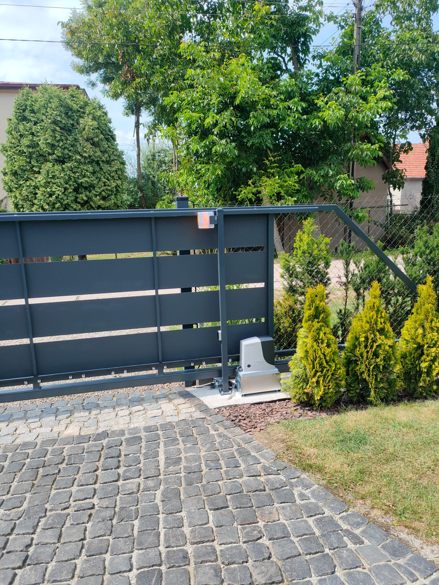 Nowoczesna, przesuwna brama wjazdowa z automatyką, widoczna z bliska na tle zieleni i kostki brukowej. Brama w kolorze grafitowym z poziomymi panelami.
