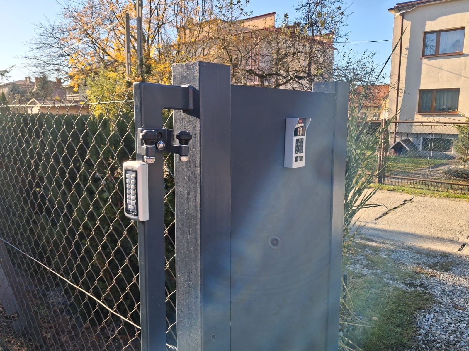 Szara brama z domofonem i klawiaturą numeryczną, widok z bliska na tle siatki ogrodzeniowej i zabudowań. Słoneczny dzień.