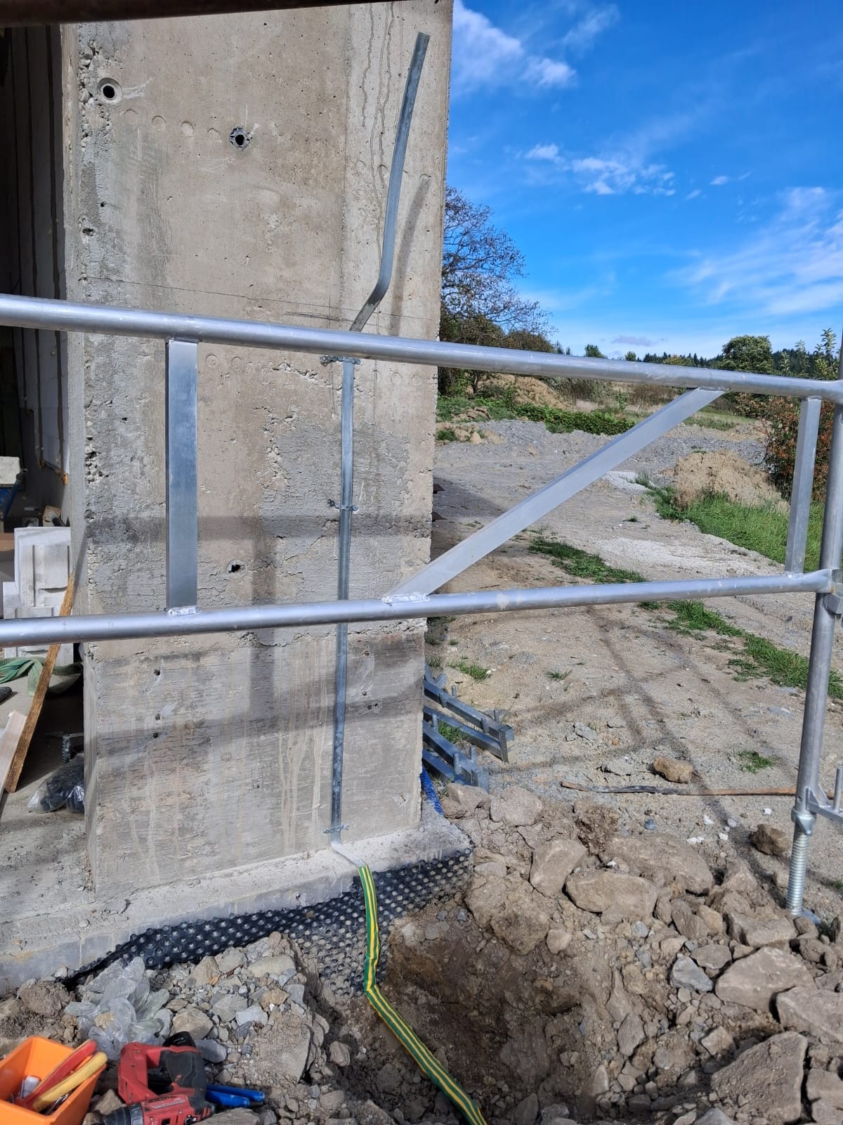 Betonowy słup z zamontowaną metalową konstrukcją, widoczny kabel w żółto-zielonej izolacji, ziemia i niebo w tle. Prace budowlane w toku.