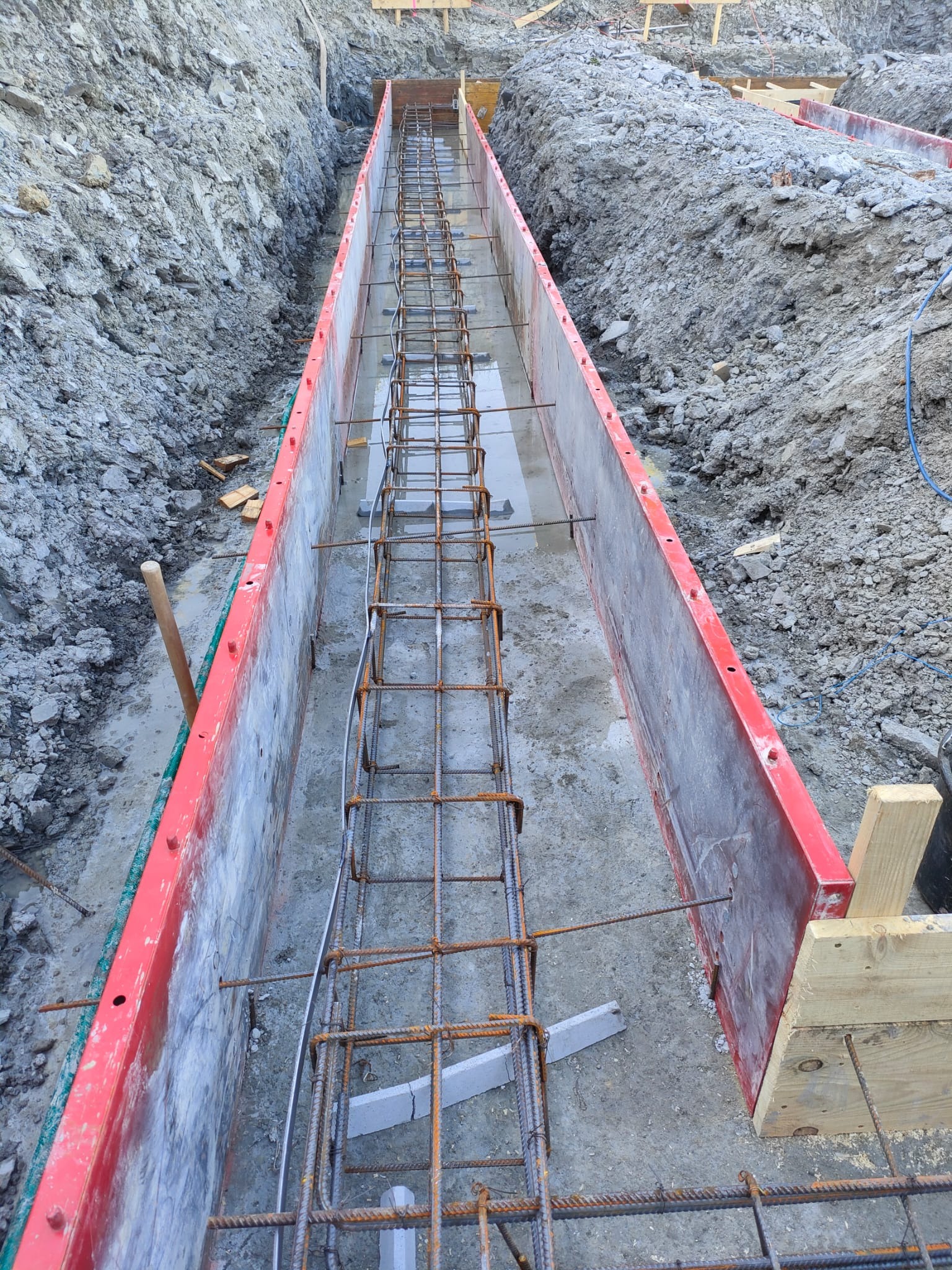 Szeroki kadr na wykop z szalunkiem i zbrojeniem fundamentów. Widoczne stalowe pręty zbrojeniowe ułożone wzdłuż wykopu, przygotowane do zalania betonem.