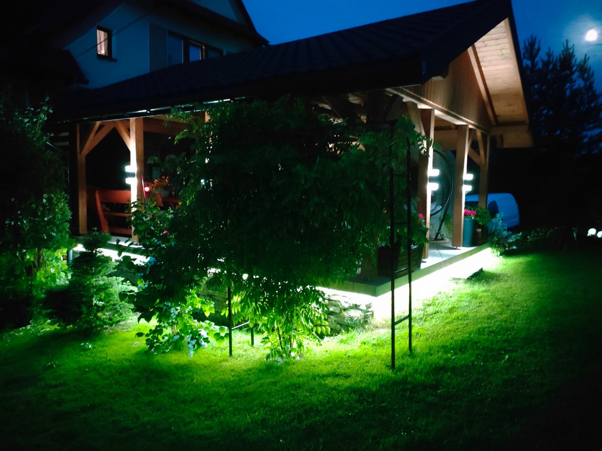 Oświetlona altana ogrodowa nocą, z podświetlonymi roślinami i elementami konstrukcyjnymi, tworząca nastrojowy klimat. Widoczne detale drewnianej konstrukcji i zielony trawnik.