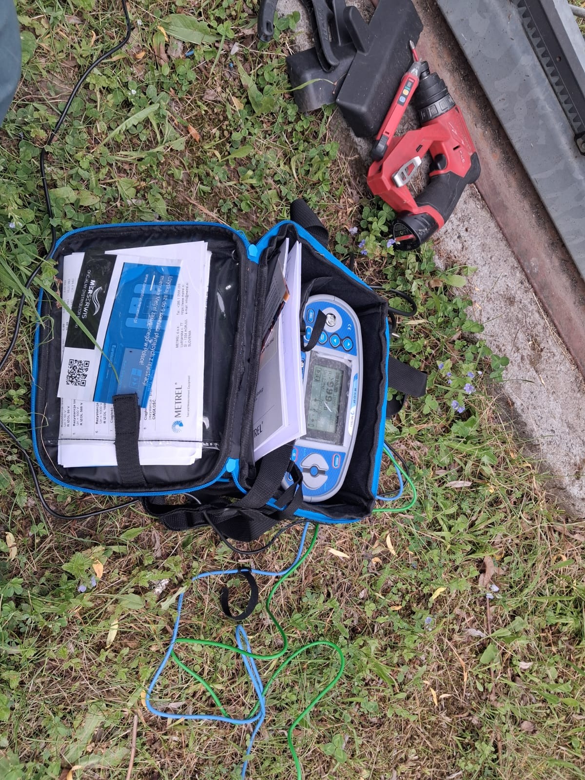 Otwarta torba z miernikiem Metrel, dokumentami i przewodami na trawie, obok wkrętarka akumulatorowa. Sprzęt do pomiarów elektrycznych w terenie.
