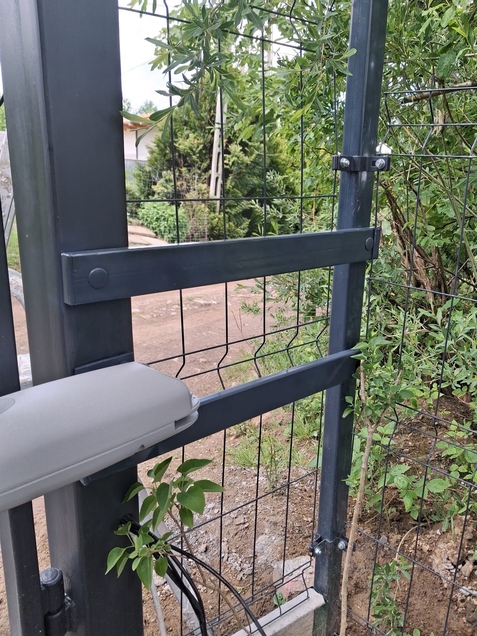 Szczegółowe ujęcie szarej bramy z siatki z zamontowanym napędem, widoczne elementy mocujące i przewody. Tło stanowi zieleń roślinności i fragment budynku.