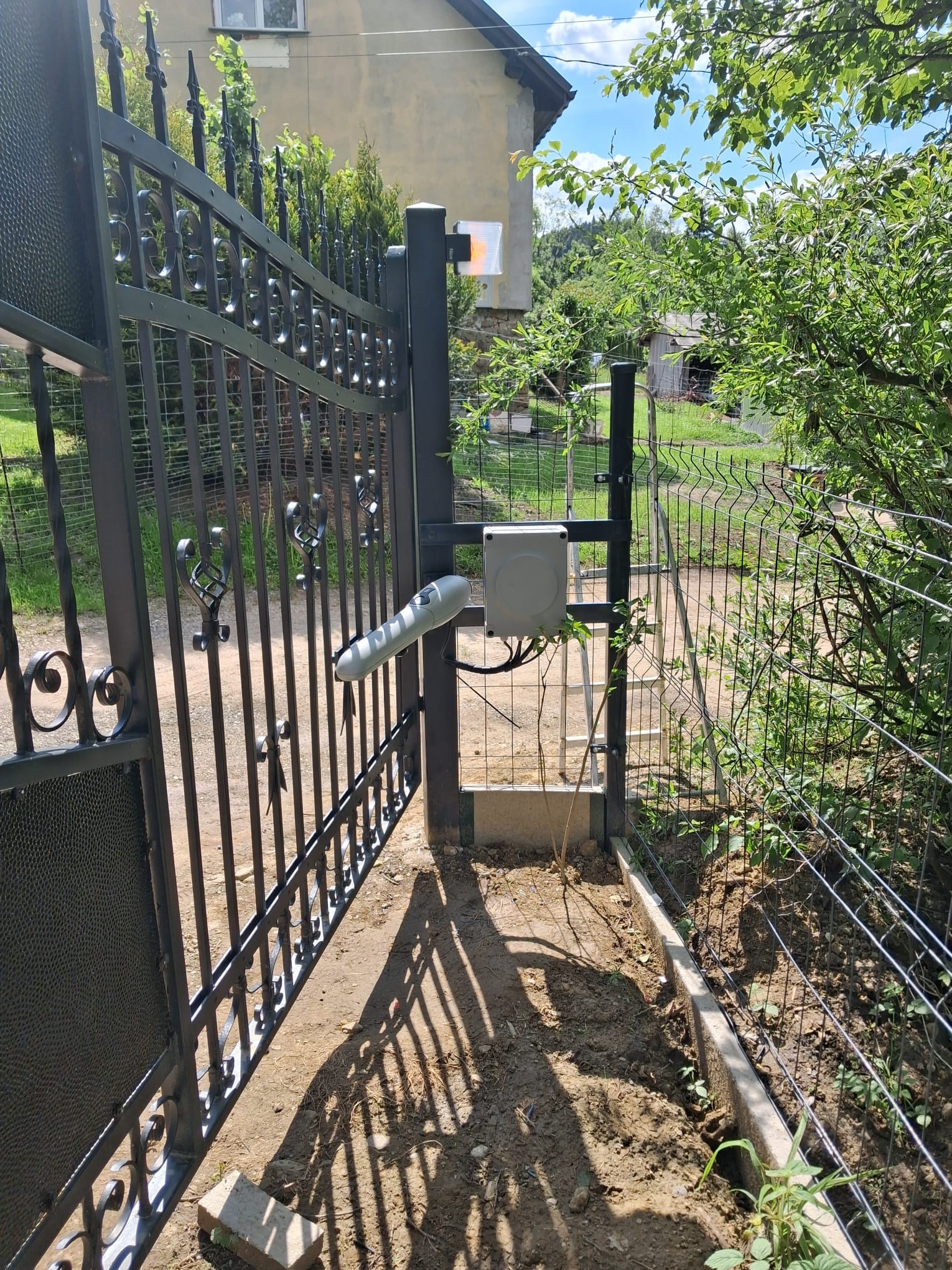 Czarna, metalowa brama z zamontowaną automatyką, widoczny siłownik i skrzynka sterująca. Tło: budynek, zieleń. Słoneczny dzień.