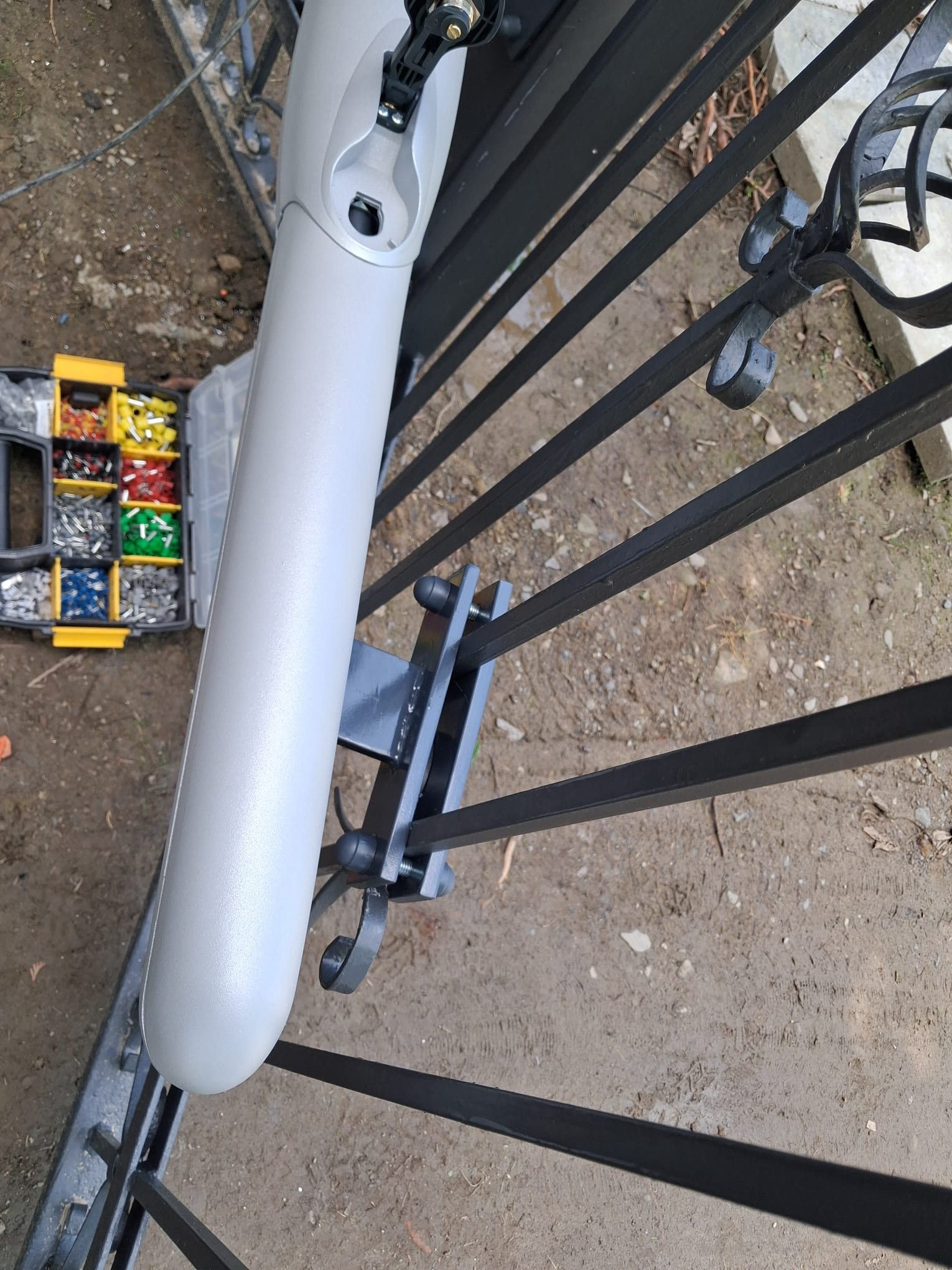 Szary siłownik zamontowany do czarnej, metalowej bramy. Obok skrzynka z konektorami. Widok z góry, na zewnątrz.