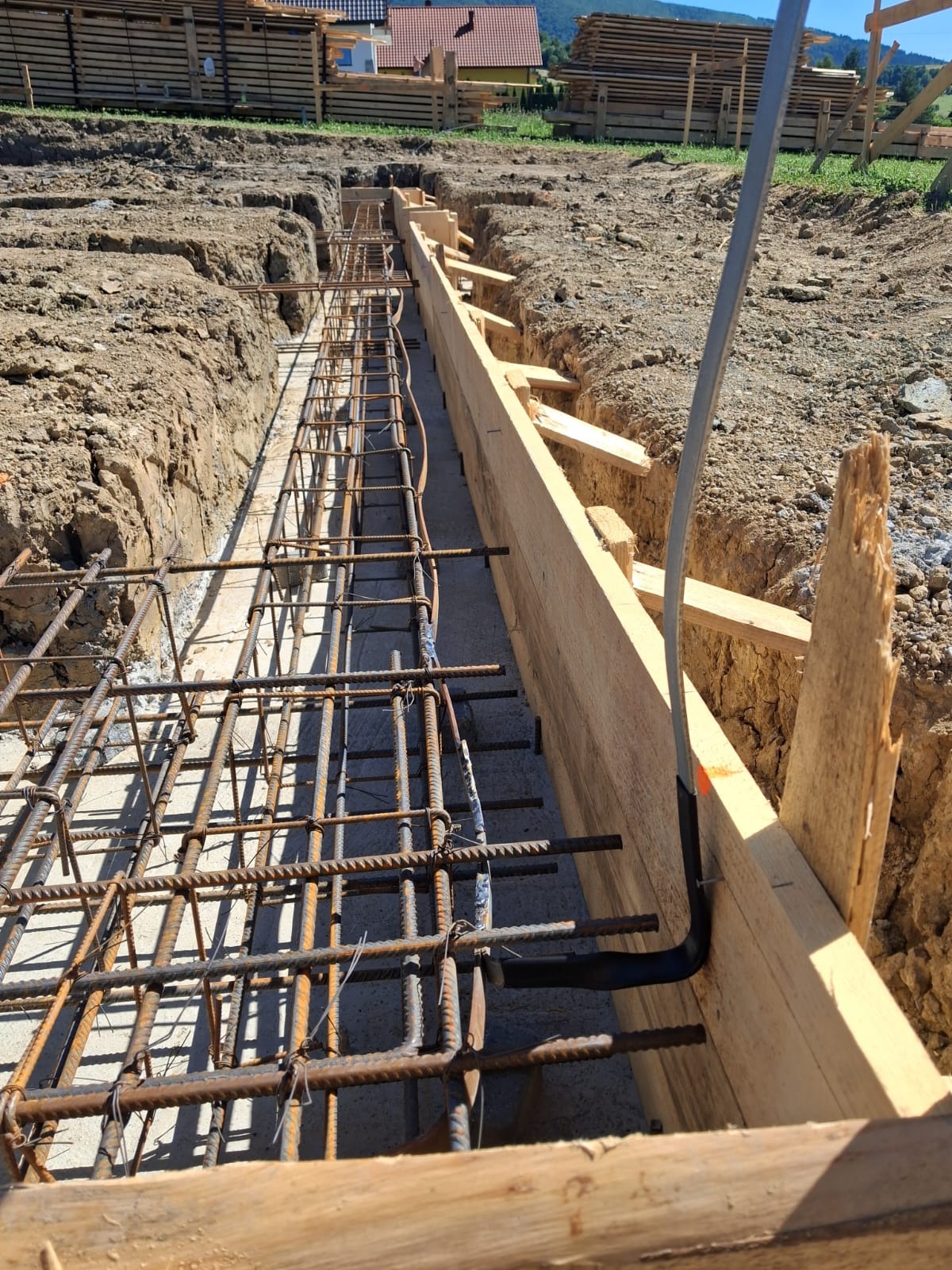 Zbrojenie fundamentów z wyprowadzonym przewodem elektrycznym w szalunku drewnianym. Widok na wykop, w tle dom w budowie i zalesione wzgórza.