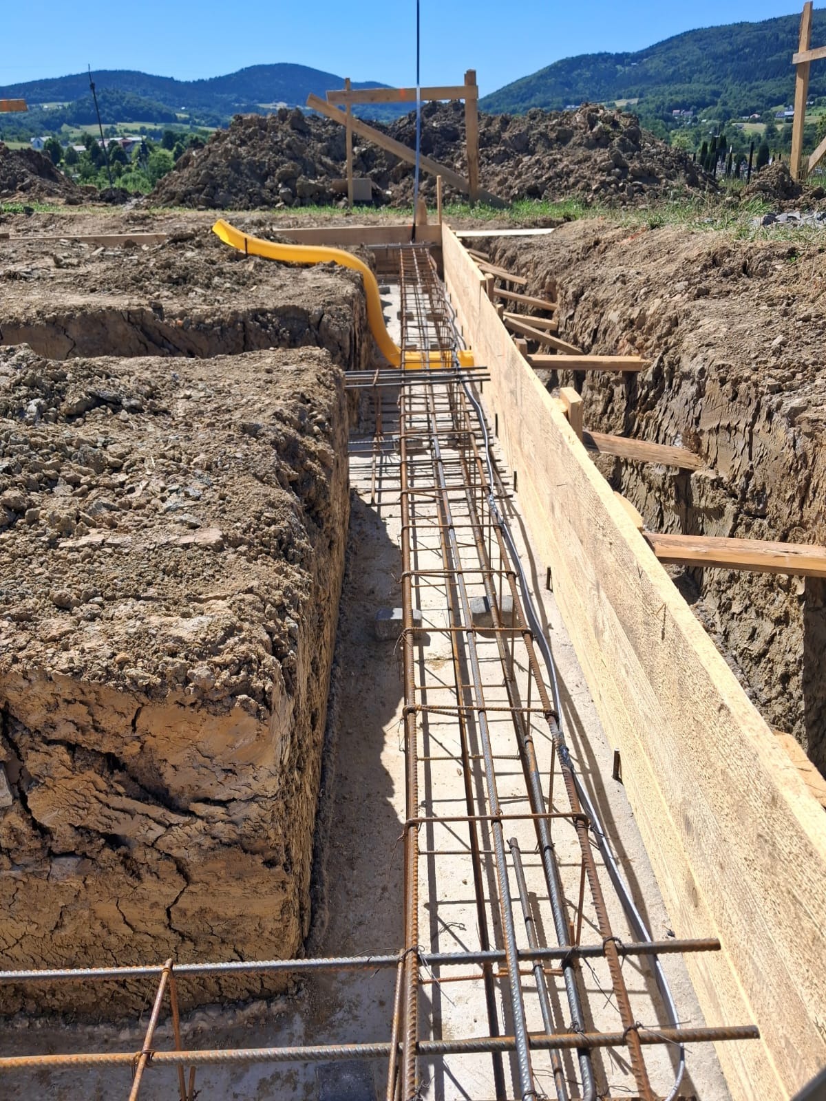 Zbrojenie fundamentów w wykopie z drewnianym szalunkiem, widoczna żółta rura instalacyjna. W tle góra ziemi i zalesione wzgórza w słoneczny dzień.