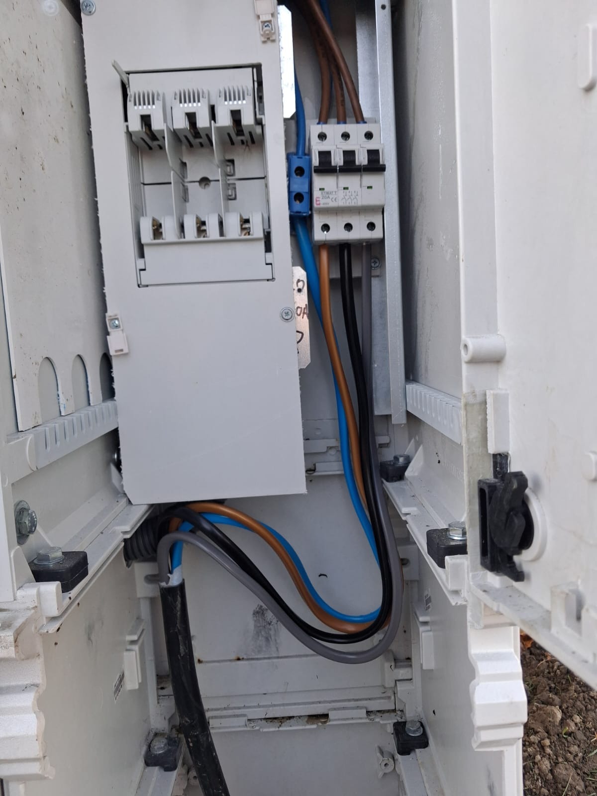 Otwarta skrzynka elektryczna z widocznymi bezpiecznikami, przewodami w kolorach: brązowym, niebieskim i czarnym. Widoczne elementy montażowe.