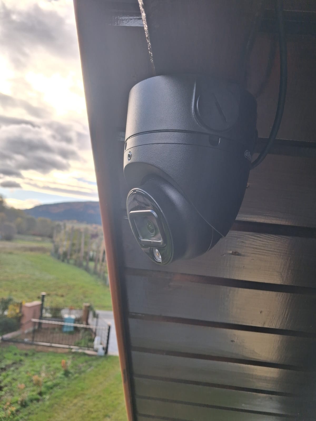 Kamera kopułkowa CCTV zamontowana na drewnianej konstrukcji, z widokiem na zielony krajobraz. Czarna obudowa kamery kontrastuje z jasnym tłem.