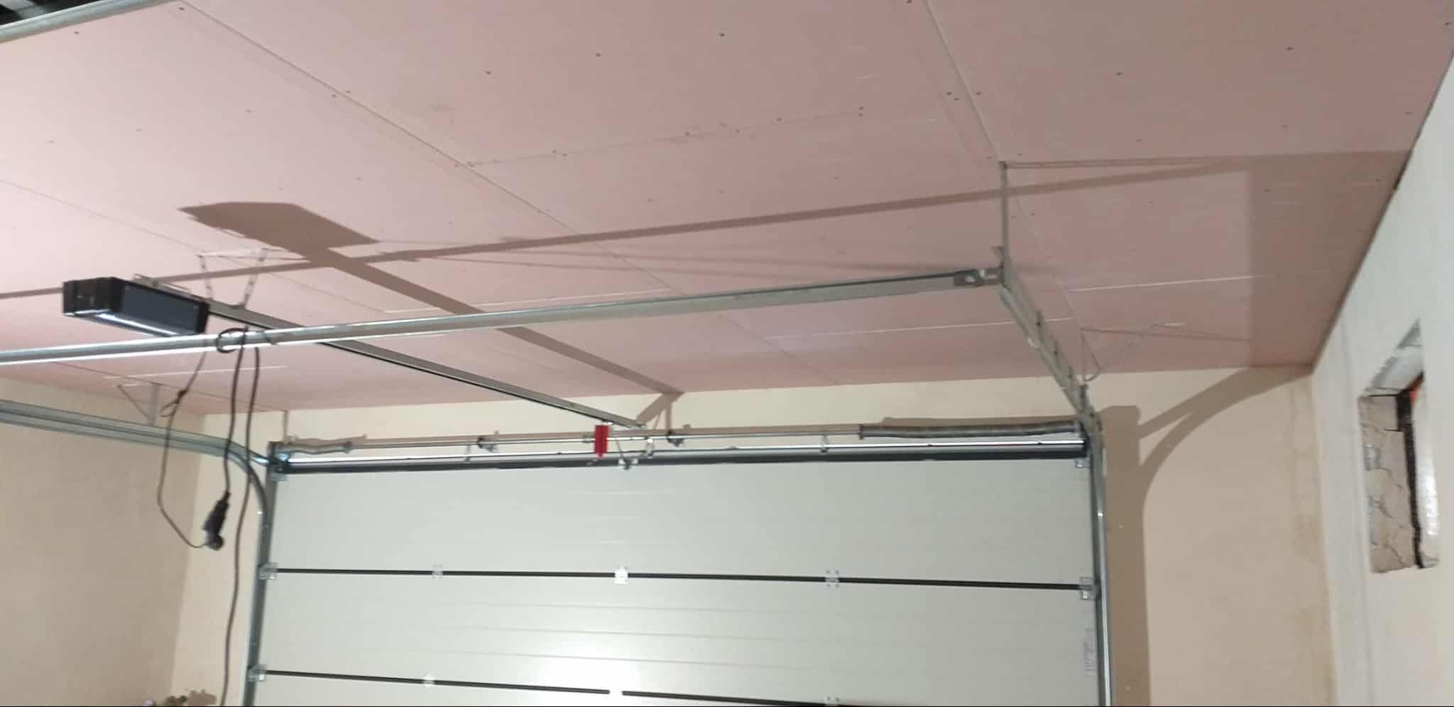 Montaż sufitu z płyt gipsowych w garażu z widocznym mechanizmem bramy segmentowej. Widoczne profile i konstrukcja nośna.