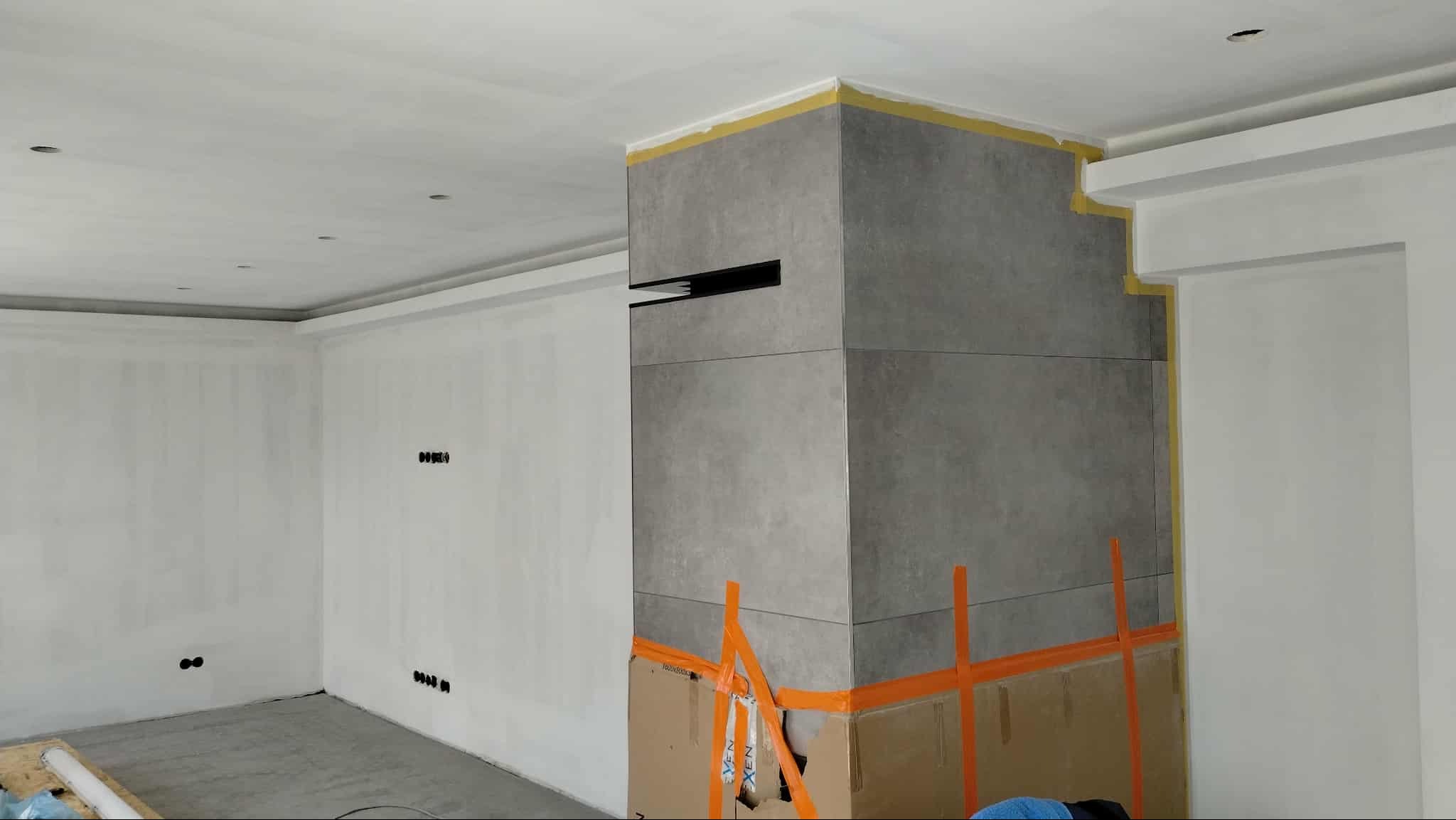 Wnętrze w trakcie wykańczania: szare płytki na ścianie zabezpieczone taśmą, surowe ściany przygotowane do malowania, widoczne puszki elektryczne. Prace wykończeniowe.