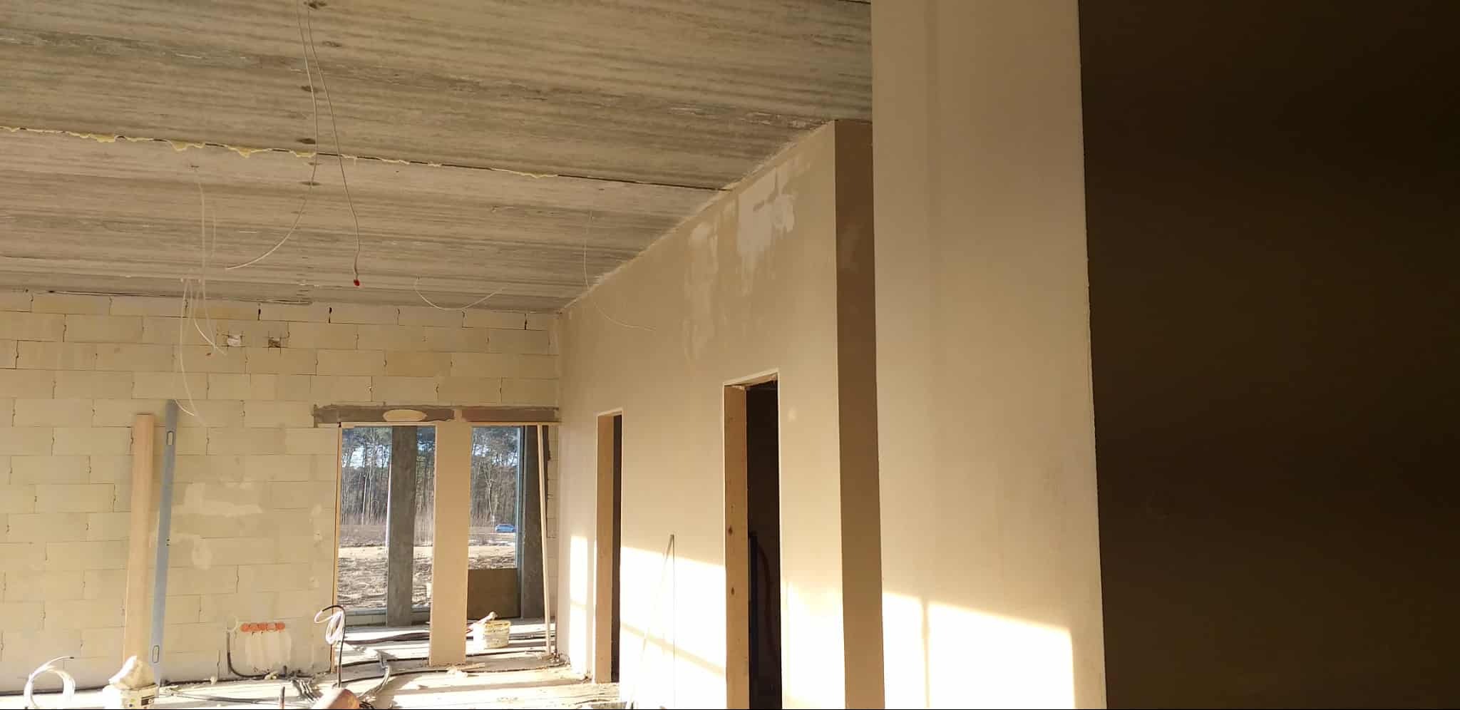 Wnętrze w trakcie prac wykończeniowych: ściany częściowo otynkowane, widoczne przewody elektryczne, surowy strop i okna z widokiem na las. Jasne, naturalne światło.