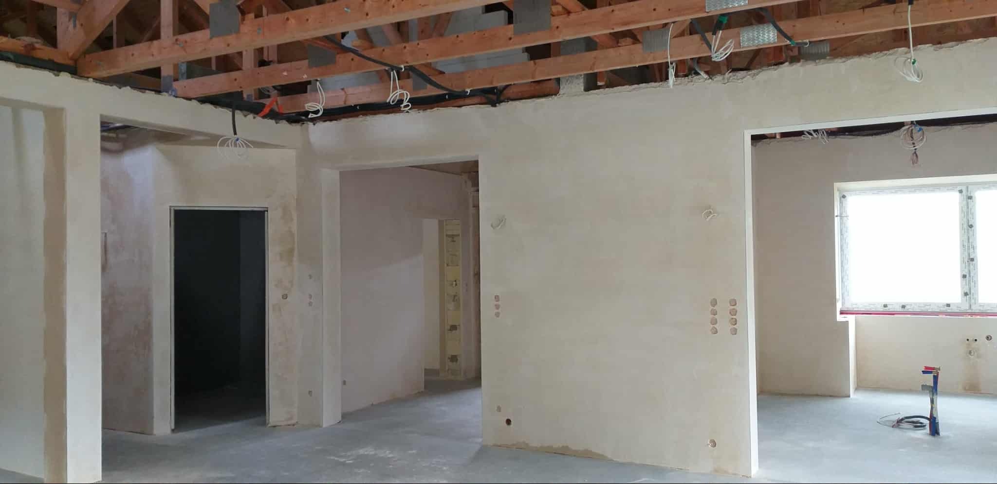 Wnętrze w trakcie tynkowania: surowy strop drewniany, świeżo otynkowane ściany z widocznymi puszkami elektrycznymi, betonowa podłoga. Widok na kilka pomieszczeń.