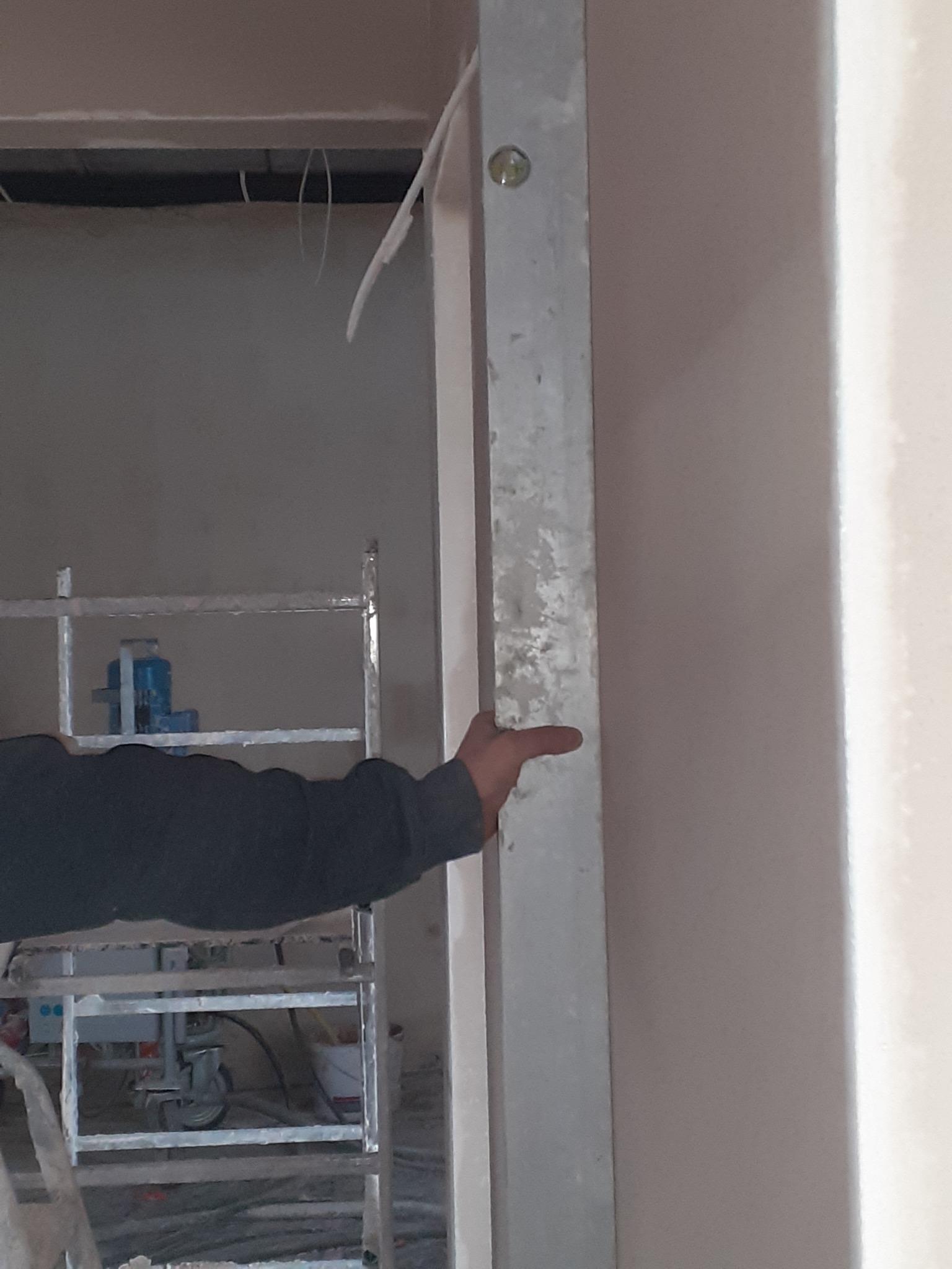 Przygotowanie ościeżnicy do tynkowania maszynowego. Widoczny fragment rusztowania i ręka pracownika. Oświetlenie naturalne, wnętrze w trakcie prac wykończeniowych.