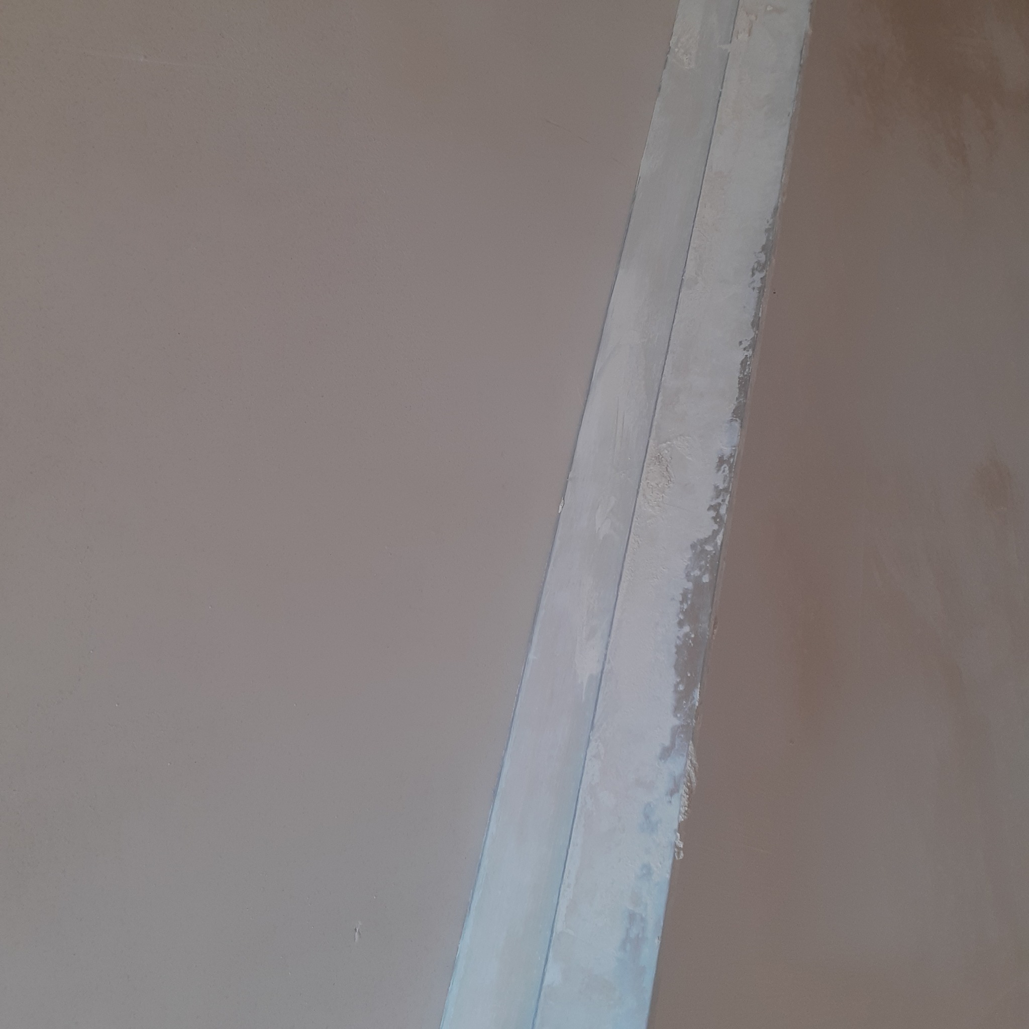 Gładka, beżowa ściana po tynkowaniu, z widoczną listwą narożnikową. Delikatna faktura tynku, neutralne oświetlenie. Minimalistyczne ujęcie detalu wykończenia.