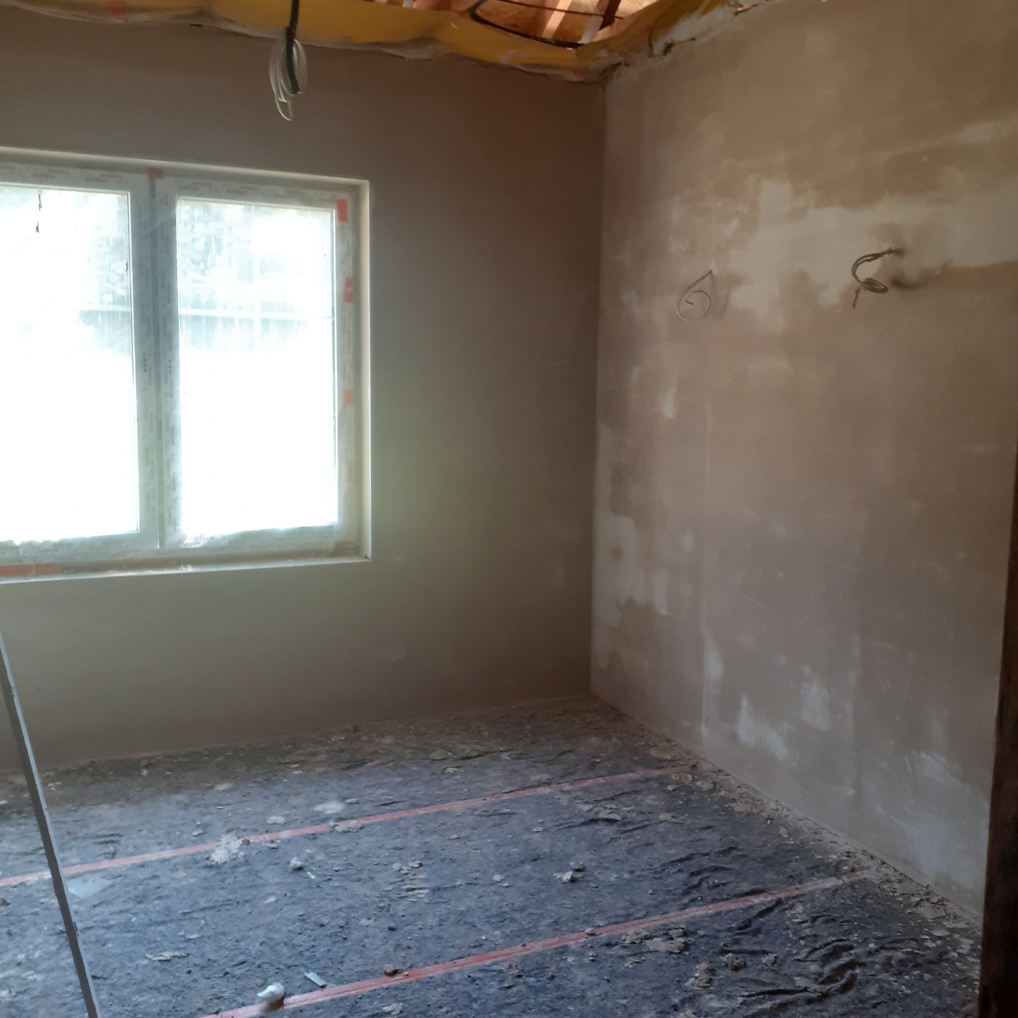 Pokój po tynkowaniu ścian, widoczne okno z parapetem, fragment podłogi z instalacją. Ściany w jasnym odcieniu szarości, sufit częściowo odsłonięty, widoczne elementy konstrukcyjne.